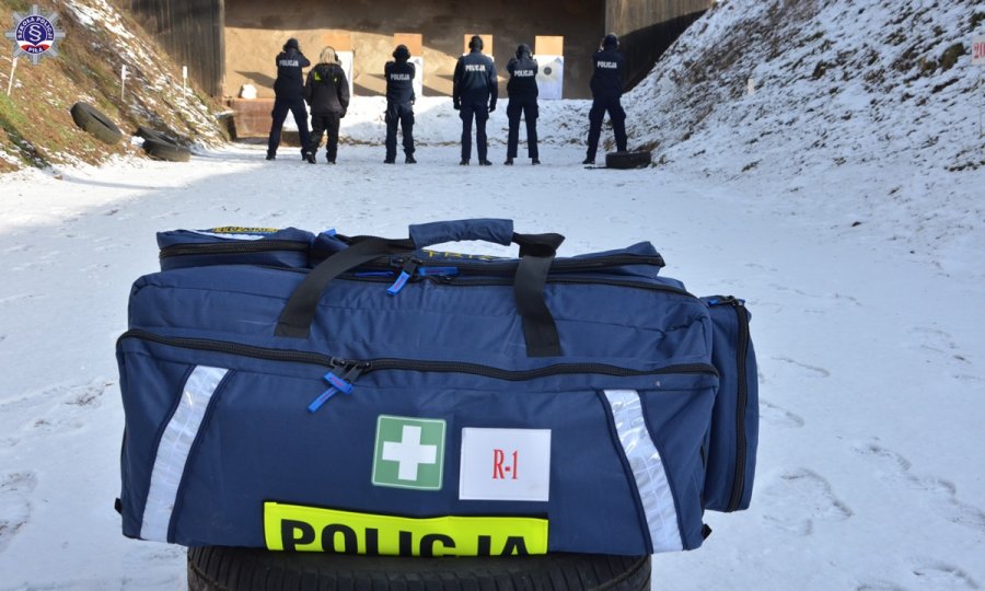 Umundurowani policjanci na osi strzeleckiej na strzelnicy. Na pierwszym planie torba medyczna koloru niebieskiego.