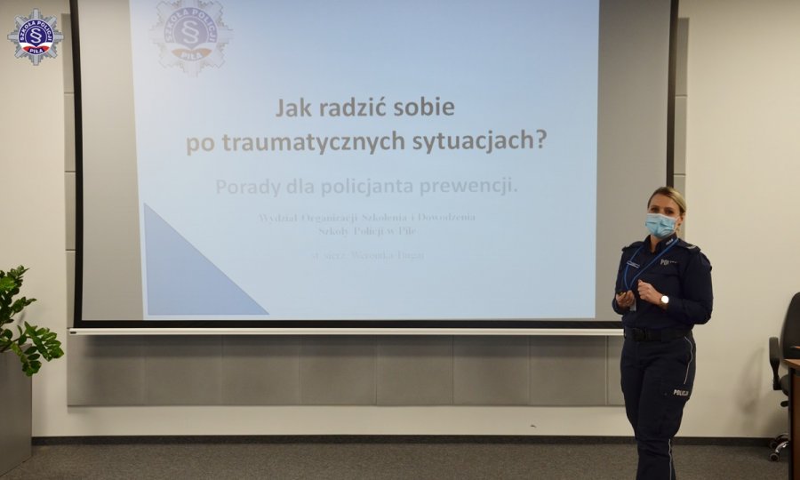 Umundurowana policjantka prowadzić wykład. W tle ekran z wyświetlaną prezentacją multimedialną zatytułowaną „Jak radzić sobie po traumatycznych sytuacjach”