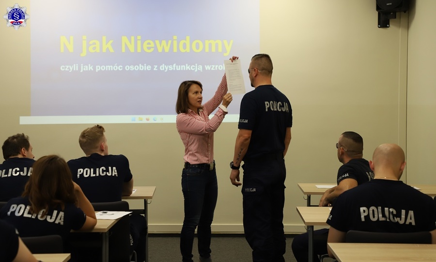 Prowadząca szkolenie dot. osób niewidomych okazuje dokument przed policjantem, który ma zasłonięte oczy. W tle policjanci.