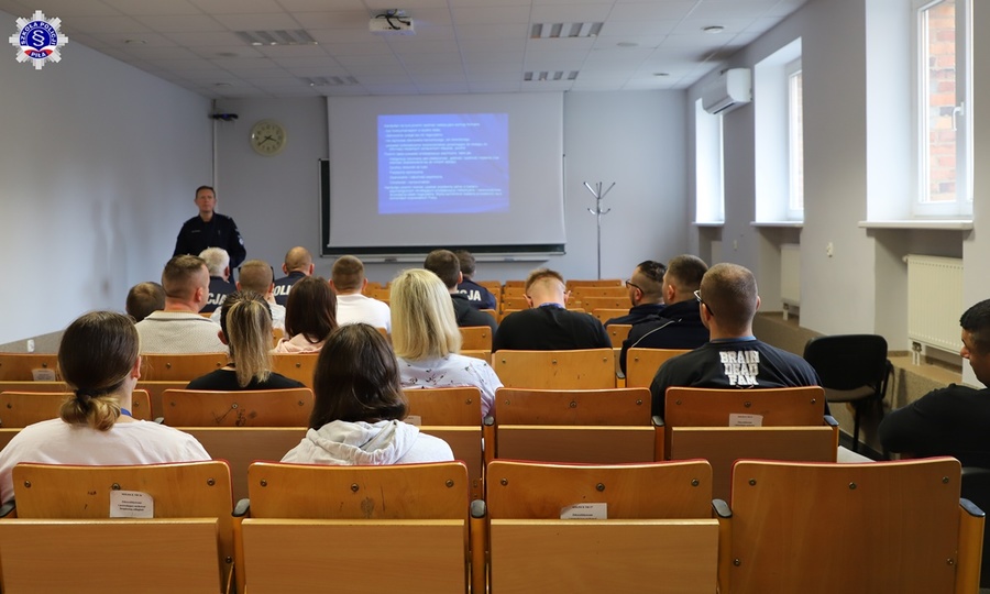 Umundurowany funkcjonariusz przemawiający na stojąco do siedzących na krzesłach nieumundurowanych funkcjonariuszy.