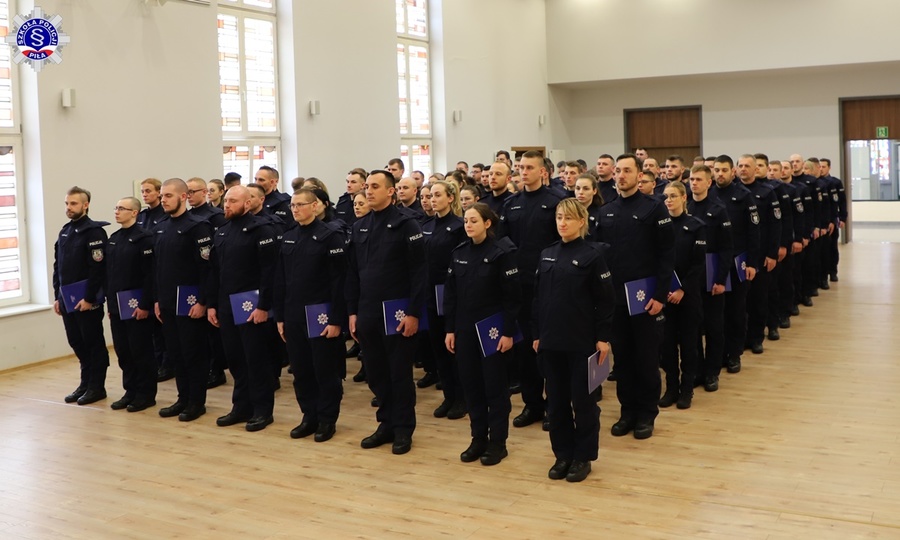 Widok z przody na ustawionych w szeregu na auli Szkoły Policji w Pile absolwentów szkolenia zawodowego podstawowego ze świadectwami podczas uroczystego zakończenia szkolenia.