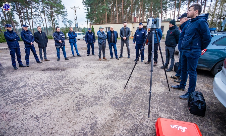 Uczestnicy warsztatów i prowadzący warsztaty przy wejściu na strzelnicę Szkoły Policji w Pile, po środku nich skaner 3D na stojaku – widok z przodu