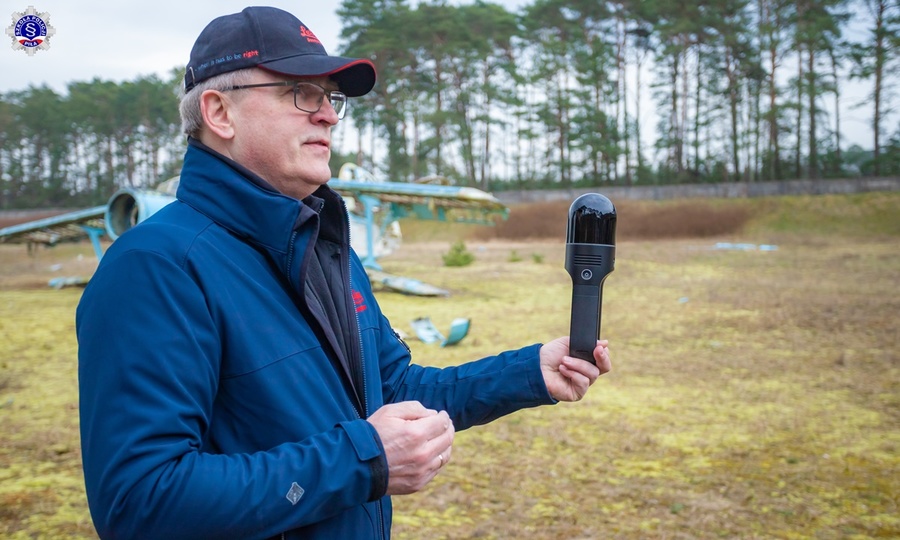 Mężczyzna trzymający w lewej dłoni skaner 3D, w tle wrak samolotu