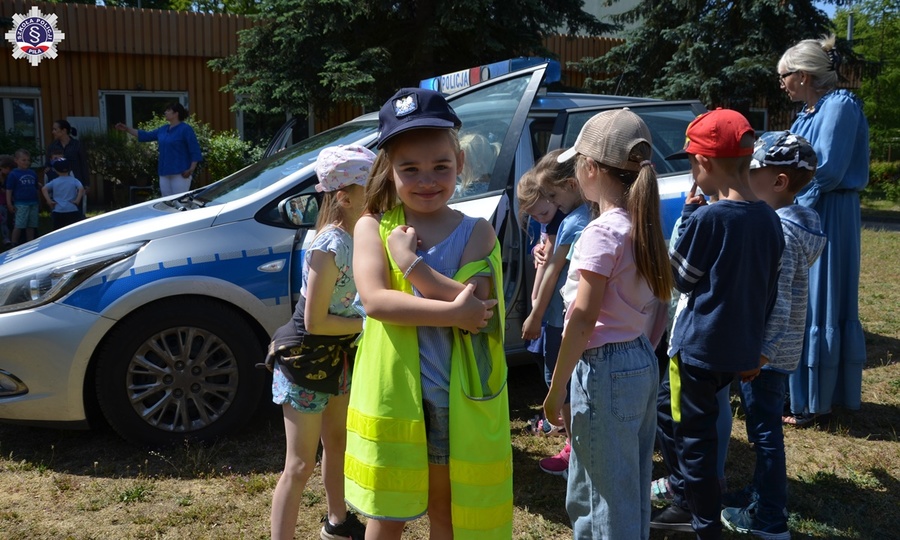 Mała dziewczynka w policyjnej czapce i kamizelce odblaskowej na tle policyjnego radiowozu. W tle inne dzieci.