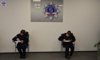 Dwie policjantki wypełniają dokumenty siedząc na krześle. W tle wiszący na ścianie zegar z logo Szkoły Policji w Pile.