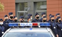 Policyjny radiowóz oznakowany, w tle stoją umundurowani policjanci w szyku.