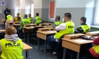 Grupa osób w kamizelkach odblaskowych w z napisem POLICJA w siedzi w sali podczas zajęć.