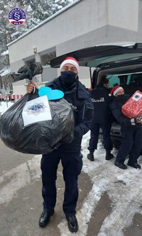 Umundurowany policjant w czapce świętego Mikołaja niesie worek z prezentami.