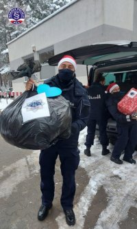 Umundurowany policjant w czapce świętego Mikołaja niesie worek z prezentami.
