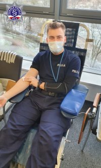 Mężczyzna w policyjnym mundurze, siedząc na fotelu trzyma się za rękę uciskając miejsce z którego pobierana była krew.