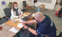 Policjant w maseczce podpisuje niezbędne dokumenty, na blacie biurka za którym siedzi pielęgniarka.