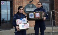Dwie umundurowane policjantki stoją przed wejściem do Szkoły Policji w Pile i  trzymają w rękach paczki.