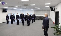 Umundurowany Policjant stoi w postawie zasadniczej, w tle grupa stoi umundurowanych policjantek i policjantów.