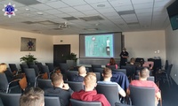 Umundurowana policjantka prowadzi wykład. Grupa osób, kobiety i mężczyźni siedzący na sali wykładowej. W tle na ekranie wyświetlana prezentacja multimedialna.