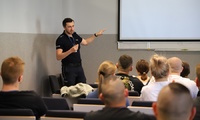 Umundurowany policjant prowadzi wykład w sali wykładowej. Grupa słuchaczy uczestniczy w zajęciach.
