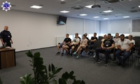 Umundurowany policjant prowadzi wykład. Grupa słuchaczy biorąca udział w zajęciach.