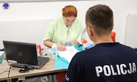 Umundurowany policjant oraz kobieta w lekarskim fartuchu siedzący przy stole rejestracyjnym dawców krwi.