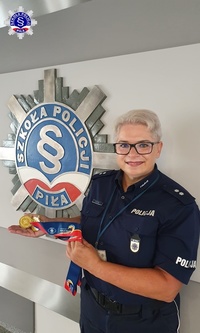 Umundurowana podkom. Małgorzata Krzyżan trzymająca w rękach swój złoty medal po powrocie z zawodów. W tle logo Szkoły Policji w Pile.