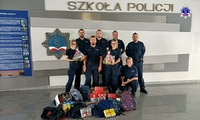 Grupa umundurowanych policjantek i policjantów na wspólnym zdjęciu na tle logo Szkoły Policji w Pile. Na pierwszym planie przygotowane i zapakowane prezenty.