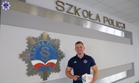 Umundurowany policjant stojący na holu szkoły, w tle logo Szkoły Policji w Pile