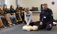 Dziecko ćwiczy masaż serca na manekinie pod okiem umundurowanego policjanta.