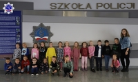 Zdjęcie grupowe przedszkolaków wraz z panią wychowawczynią przed logo Szkoły Policji w Pile w budynku głównym Szkoły.