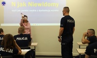 Prowadząca szkolenie dot. osób niewidomych trzyma w górze rękę przed policjantem, który ma zasłonięte oczy, w tle inni policjanci.