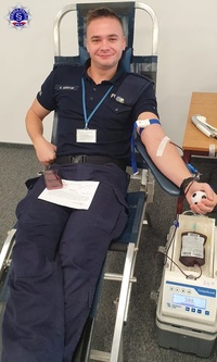 Mężczyzna w policyjnym mundurze, siedząc na fotelu oddaje krew i uśmiecha się do obiektywu.