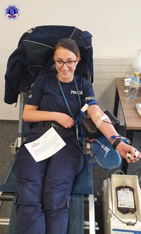 Kobieta w okularach w policyjnym mundurze, siedząc na fotelu oddaje krew i uśmiecha się do obiektywu.