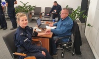 Policjantka siedzi na fotelu obok mężczyzny siedzącego za biurkiem wypełniający dokumenty. W tle widać policjantów oczekujących na badanie.