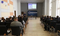 Grupa osób w Auli Szkoły. W tle umundurowana policjantka prowadząca wykład. Widoczny ekran, na którym wyświetlana jest prezentacja multimedialna.