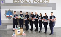 Zdjęcie grupowe na tle logo Szkoły Policji w Pile wolontariuszy szkolnego samorządu, którzy trzymają w ręku prezenty dla podopiecznych szpitala. Przed nimi stoi pudełko wypełnione prezentami.
