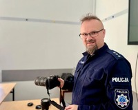 Komisarz Marcin Mańczak trzyma aparat fotograficzny w prawej dłoni, uśmiecha się.