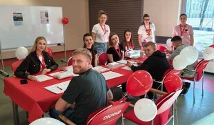 wolontariusze oraz potencjalni dawcy siedzą przy stoliku rejestracyjnym i uśmiechają się do zdjęcia