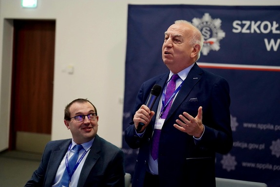 Na pierwszym planie po prawej stronie stoi sędzia Kozielewicz w granatowym garniturze, jasnej
koszuli i fioletowym krawacie, który przemawia do trzymanego w prawej dłoni mikrofonu i
gestykuluje lewą ręką. Po lewej stronie siedzi uśmiechnięty prof. Jóźwiak w okularach, ciemnym
garniturze i niebieskim krawacie, spoglądający w stronę mówcy. Obaj uczestnicy mają założone
niebieskie smycze z identyfikatorami i napisem „Szkoła Policji w Pile”. W tle po prawej stronie
znajduje się granatowy baner z logotypem, fragmentem nazwy oraz adresem strony
internetowej Szkoły Policji w Pile.