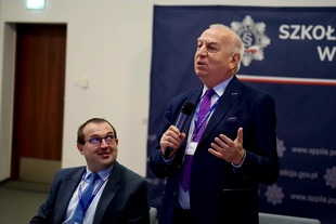 Na pierwszym planie po prawej stronie stoi sędzia Kozielewicz w granatowym garniturze, jasnej
koszuli i fioletowym krawacie, który przemawia do trzymanego w prawej dłoni mikrofonu i
gestykuluje lewą ręką. Po lewej stronie siedzi uśmiechnięty prof. Jóźwiak w okularach, ciemnym
garniturze i niebieskim krawacie, spoglądający w stronę mówcy. Obaj uczestnicy mają założone
niebieskie smycze z identyfikatorami i napisem „Szkoła Policji w Pile”. W tle po prawej stronie
znajduje się granatowy baner z logotypem, fragmentem nazwy oraz adresem strony
internetowej Szkoły Policji w Pile.